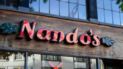 Getty Images A Nandos sign with red writing and a brown wooden background. The sign sits between two glass windows 