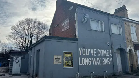 An end terrace house painted grey, with signage reading The New Adelphi Club and large white lettering on the wall saying “You’ve come a long way, baby”. A bicycle stand and an arched doorway are situated on the front of the building. Trees and a cloudy sky are visible in the background.
