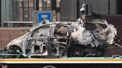 Niall Carson/PA Wire The burnt wreckage of a white car which exploded outside Dunmurry Police Station on Saturday. The bodywork is very badly damage, with the metal twisted out of shape in many places. All of the windows have been blown out and parts of the doors are missing. The car has been loaded on the back of a low loader to be towed away.