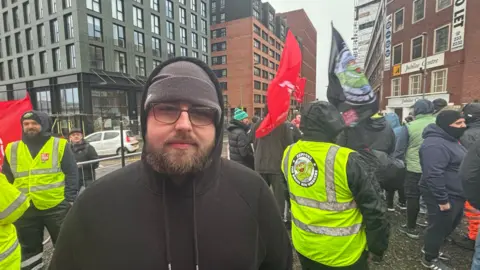 Agency worker Luke Graham wears a black hoodie with the hood up. He also has a grey woolly hat and glasses. Other workers can be seen behind him wearing bright yellow hi-vis jackets. Some are carrying flags. 
