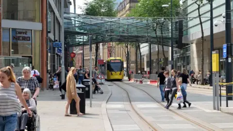 Manchester city centre on a busy day, with pedestrians seen passing over tramway close the the Arndale shopping centre, with a yellow bee network dram seen in the distance, and a public walkway connecting two buildings pictured above.
