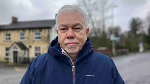An older man with grey hair stands on a country lane. He is wearing a navy coat. There is a stone house behind him and some trees.