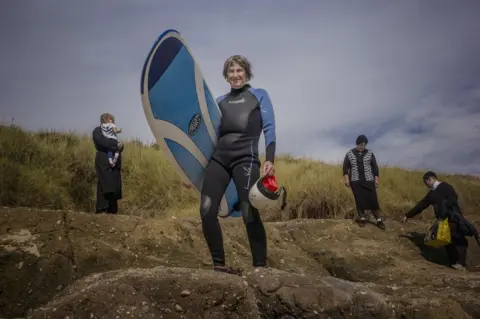Sarah Lee A woman holds a surfboard whilst an Orthodox Jewish family gather behind her