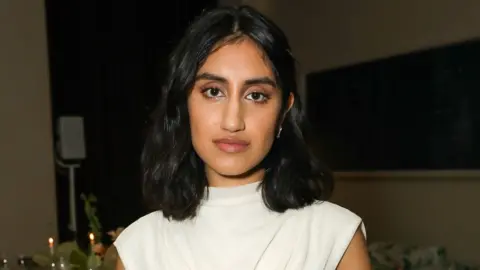 Getty Images Ambika Mod. Ambika is a 28-year-old British Asian woman with shoulder length black hair which she wears loose and large brown eyes. She looks at the camera with a serious expression. She wears a high-necked short sleeved white dress, matched with a long gold chain. She's pictured inside at night at what appears to be a dinner party