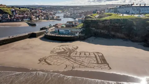 Getty Images A giant sand portrait by WaterAid and artists Sand In Your Eye of Ansha, a 12-year-old from Frat in Ethiopia adorns Whitby Beach on March 15, 2021 in Whitby, England. Ahead of World Water Day on 22 March WaterAid created the portrait in the sand on Whitby Beach of a child carrying water next to the rising tide, to highlight the impact climate change is having on people’s access to water. The 60-metre wide artwork depicts Ansha from Frat in Ethiopia, who spends hours each day walking to and collecting dirty water from a river. After only an hour Ansha’s image was washed away by the rising tide – highlighting how excess rainfall and rising sea levels can lead to flooding, contaminating water and endangering lives.