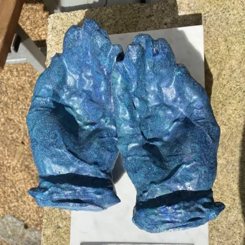 The Nurse's Hands sculpture viewed from above. It is a pair of cupped hands formed out of a blue material. There is a silver-coloured plate below it with the inscription: The Nurse's Hands 2024 Recycled PPE By Kurt Jackson Dedicated to those who care and those we lost