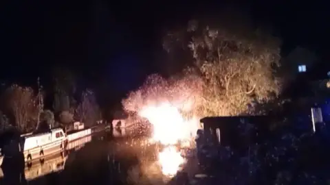 Tony Gosling A large ball of flames on the river, illuminating a nearby tree, sparks can be seen shooting out of the flames, there are two other cruiser boats moored up near the blaze.