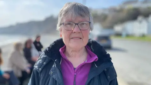 Marinda Faragher is wearing a purple fleece top under a dark blue coat. She had short grey hair and is wearing glasses. Laxey promenade is behind her, with people sitting on a wall to the left. It is a sunny day.