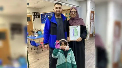 Akeel, Hannaa and Sara in Al Ashraf Primary School. Sara is wearing a warm coat, pink earmuffs and has a hand puppet. Hannaa is holding a drawing done by Sara.