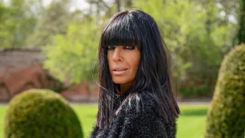 BBC/Studio Lambert/Euan Cherry Claudia Winkleman, with long black hair past her shoulders, looks sideways into the camera with a slight grin while standing among green trees in a garden.