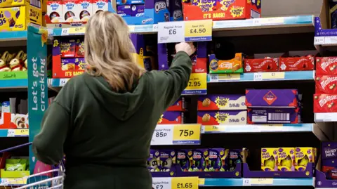 EPA A woman holding a basket in one arm and reaching for an Easter egg with the other at a shelve stocked with lots of chocolate eggs in a shop.