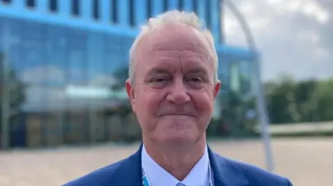 A man looking at the camera and smiling. He is wearing a blue suit, blue shirt and blue tie in front of a large glass building.