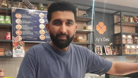 LDRS A man with short black hair and a beard and grey T-shirt is smiling while standing in a deli. Cakes can be seen behind him.