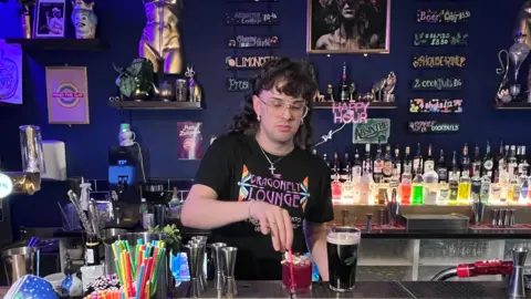 A man with long hair wearing glasses and a crucifix necklace wearing a black T-shirt stirring a red drink in a tumbler behind a colourful bar.