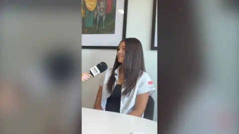 A young girl with brown hair sat in a chair. She's facing a reporter who is holding a mic to her face.