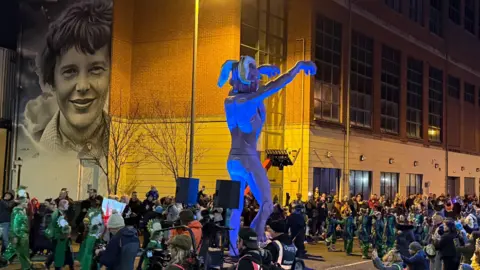 Shows large crowds in the street as the Halloween parade passes with a large puppet in the middle on wheels and a mural of Amelia Earhart in the background