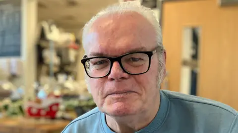 Neil with white, thinning hair, black-rimmed glasses, wearing a light blue sweatshirt, sits inside a café. 
