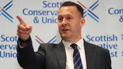 PA Media Russell Findlay, with dark hair and a dark suit, pointing in front of Scottish Conservative signage during a press conference 