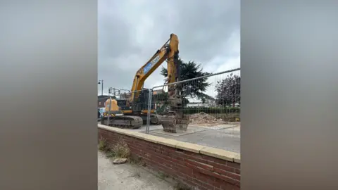 Imogen Lee/BBC A yellow digger behind a metal fence. Beyond it is a pile of rubble.