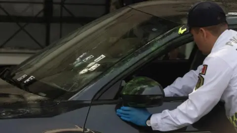 Reuters A police officer wearing blue plastic gloves and a white uniform shirt examines the steering wheel of the car of the mayor's private secretary. Bullet holes can be seen in the car's windscreen. Signs with numbers have been attached to the screen to mark the entry holes.