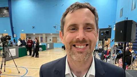 A man with short hair and facial hair standing in a sport halls smiling directly into the camera