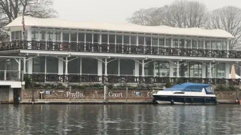 Phyllis Court pavillion is right next to the river Thames. It is a glazed building with white supports. In the foreground is the river with a boat moored alongside the building. There is a large white sign reading Phyllis Court on a brick wall in front of the building.