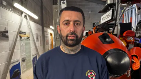 Crispin Rolfe/BBC Carl Handley pictured inside a building standing next to a lifeboat. He is wearing a blue sweat shirt featuring a Hornsea Inshore Rescue emblem. He has short dark hair and a dark beard. 