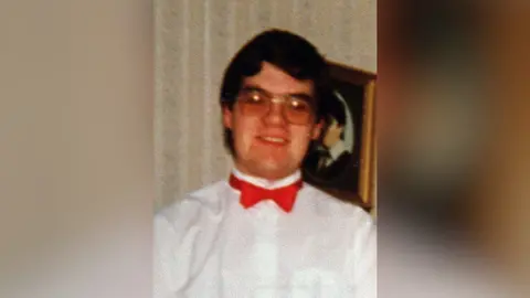 Family Handout Young man wearing glasses, a white shirt and red bowtie smiling at the camera. He is standing in front of a framed photograph handing on a wall.