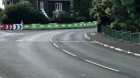 The road at Cruickshanks, which sees the road bent towards May Hill. There are crash barriers on the outside of the bend and pavement on the inside.