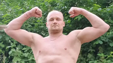 A topless man posing with his arms up showing off his muscles while standing in front of a green bush