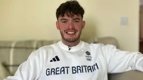 Jake Zuckerman/BBC Ed Lowe sitting on a sofa wearing his Great Britain jumper, proudly looking at the camera