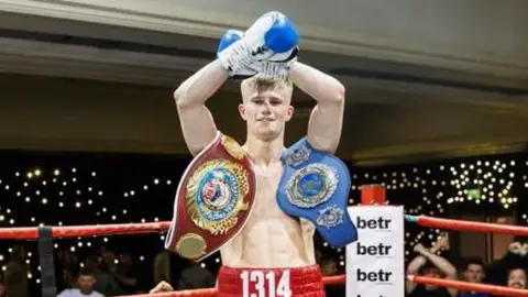 Kynoch Boxing Scotland Colin Cairney is in a boxing ring with people surrounding it. He is wearing blue boxing gloves and holding his hands aloft, crossed over. He has no top on and two  boxing championship belts slung across each shoulder. He has red boxing shorts on with 1314 on the waistband.