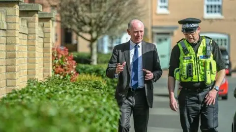 Darryl Preston wearing a blue tie and black suit, walking and talking along the streets of Peterborough with a uniformed police officer. 