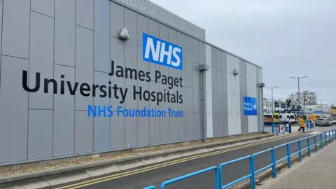 Andrew Turner/BBC The outside of the James Paget University Hospital building. The grey outside of the building has black and blue writing on it which says NHS James Paget University Hospitals NHS Foundation Trust.