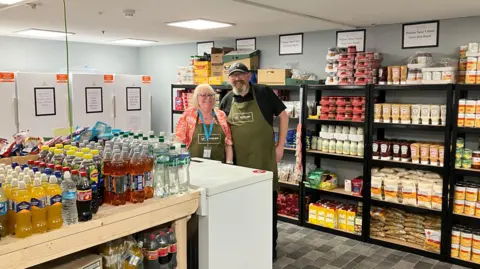 A man and woman in green aprons are standing in front of shelves of food. There is a freezer in front of them, and a table full of bottles of soft drink