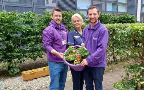 Stroke Association Shows two men wearning purple hooded tops and holding a flower pot with a nurse in a blue top and black trousers in the middle.