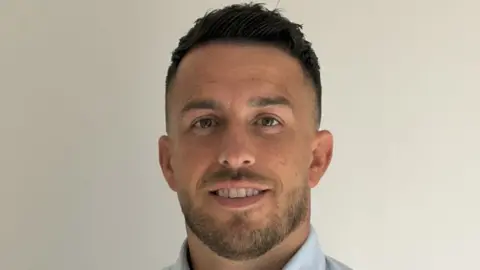 A headshot of Nicky Kingsnorth, who has black hair, short at the sides, and a close-cropped black beard. He is smiling. His light blue shirt collar can just be seen.