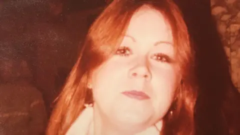 Maggie Montgomery An old photograph of a young woman. The colours are washed out and look orangey/red. She has long hair, parted in the middle and is staring at the camera with a small smile on her face. 