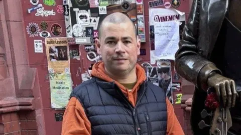 Steve Mason A man wearing blue and orange, with a shaved head, is looking at the camera. Small posters are on a burgundy-coloured wall behind him.