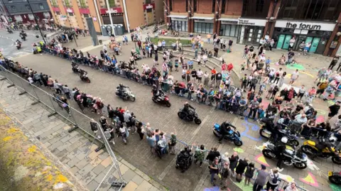 A procession of motorcyclists ride down Barrow high street. There are many people lined behind barriers in support of the event.