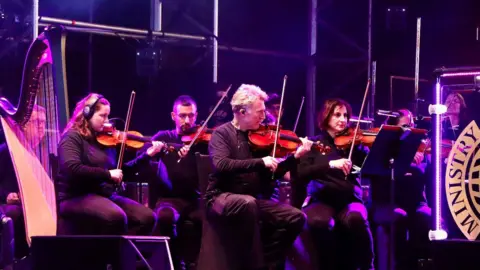 Five orchestral performers sitting down and playing violins, with one on the far left playing a harp. They are all wearing black and the stage is lit up with a purple hue