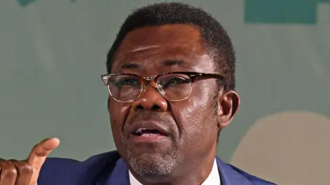 A close up shot of Veron Mosengo-Omba talking. He wears glasses and a blue suit.