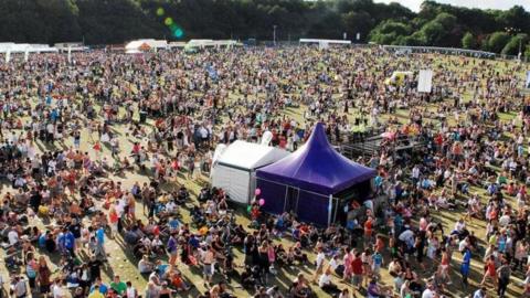 Hundreds of people sat on a grass field with a large tent in the foreground