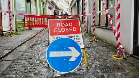 A close up of a 'road close' sign, behind a blue sign with a white arrow pointing right. They are on a cobbled road in the middle of the street.
