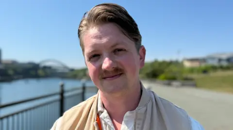 John Lee Taggart wearing a white buttoned shirt and a beige gilet style jacket stands on the Sunderland Riverside with the city's various bridges in the background. It is a sunny day.