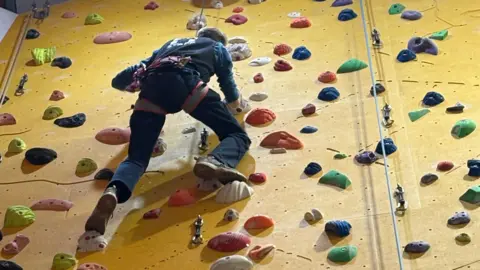 A climber is seen scaling a yellow indoor climbing wall. There are different coloured hand holds on the wall and ropes dangling down from the top.
