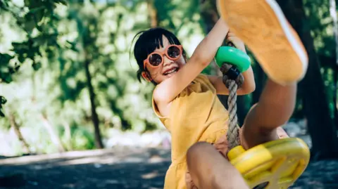 A young girl with sunglasses swings on a swing towards the camera smiling widely. 