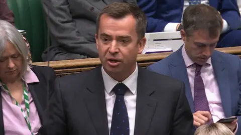 ParliamentTV Calum Miller standing up talking in parliament. He has short brown hair and is wearing a dark blazer and tie over a white shirt.
