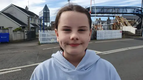 A 10-year-old girl, with brown hair tied back, wearing a light blue hoodie. She is standing in front of a road and there are houses and a bridge visible behind her. 