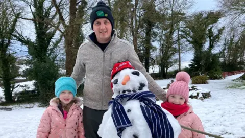 A man in a navy hat with a green crest and grey zip up top stands with two little girls in pink coats and a snow man in front of them